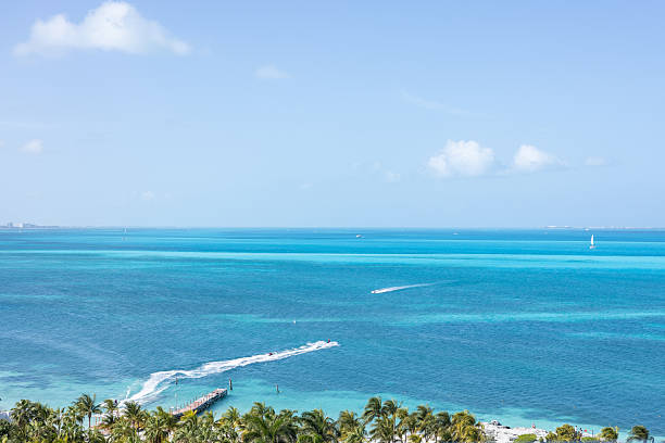 This tropical scene in Cancun showcases a pristine beach lined with soft sand, framed by palm trees and distant hotels under a clear blue sky. The vibrant turquoise waters are dotted with boats.