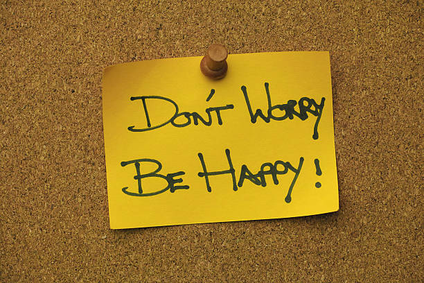 The words 'DON'T WORRY, BE HAPPY !' written in a black marker pen on yellow note paper, pinned by a wooden pin to a cork message board. Conceptual motivational and inspirational quote message relating to being content, relaxed, and carefree, etc.
