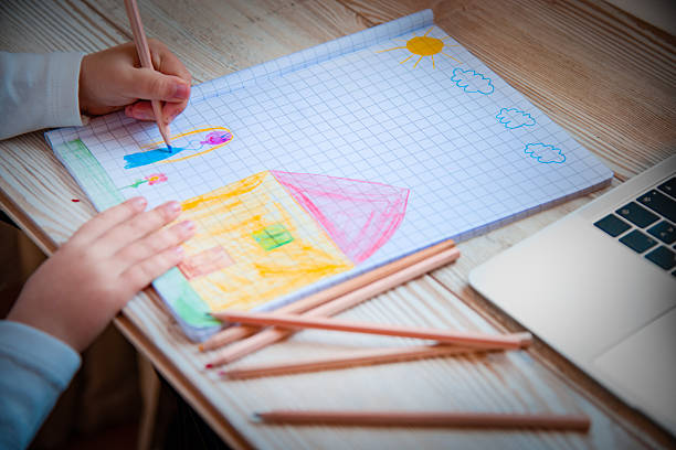 Close-up of a child drawing with colored pencils on a squared notebook. The drawing shows a colorful house, a person, the sun, and clouds. Concept of childhood creativity, early education, homeschooling, art learning, imagination, and school activities.