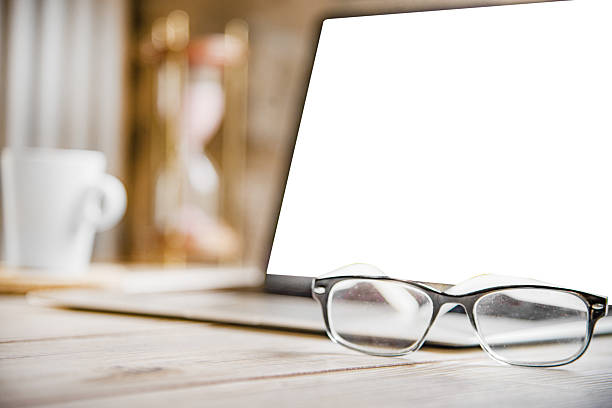 Minimalist workspace with eyeglasses placed in front of a laptop with blank white screen on a wooden desk. Cozy home office setup with coffee cup and hourglass in the background. Perfect for business, remote work, freelance, productivity, and online communication concepts.