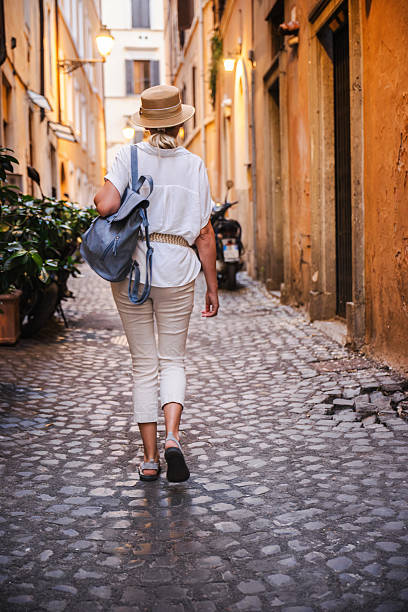 istock Middle-aged woman in summer outfit and straw hat carrying a backpack while strolling through a narrow Roman alley with warm evening light on old walls 2237658365