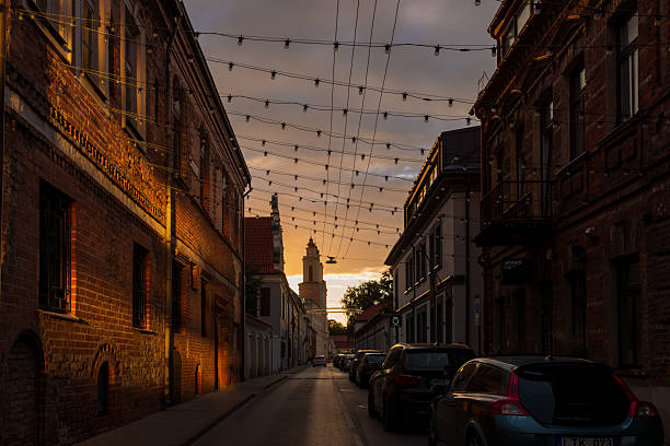 Kaunas, Lithuania – August 22, 2025: A narrow street lined with red brick façades glows in warm sunset tones. Hanging festive lights stretch above, leading the eye to a distant church tower. The mix of old walls and evening light evokes historic charm.