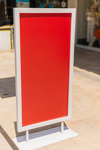 istock A tall sign with a bright red background is placed on the sidewalk. It stands empty, ready for announcements or promotions. The setting is a lively urban area on a sunny day 2237685914