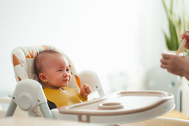 Asian baby sitting in high chair feeling upset and crying while mother feeding with spoon.