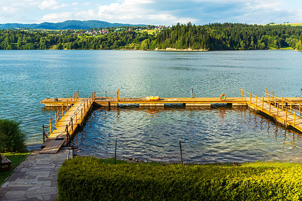 A wooden pier stretches across the reflective waters of czorsztyn lake, with distant green hills and small houses visible on the horizon. The view conveys summer leisure, rural charm, and the peaceful atmosphere of the mountain region.