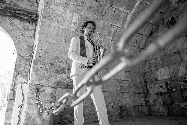 A solo saxophonist in a pale suit plays amid rough stone walls, creating a moody, atmospheric jazz moment. The foreground chain adds depth while the monochrome scene emphasizes texture and mood.