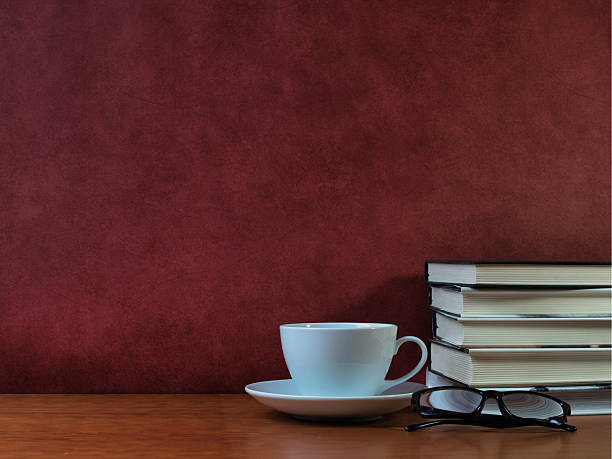 Coffee Break Study Desk Concept - White cup and saucer, stacked books, pencils, and reading glasses on a wooden desk. Professional and academic theme with generous copy space.
