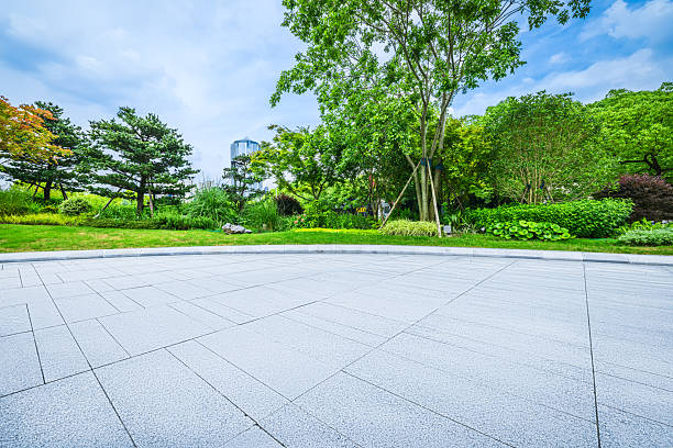 istock Empty stone tile ground in a city park with lush green trees and a lawn under a cloudy blue sky. 2237714932
