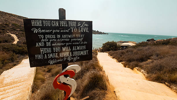 A winding coastal path leads to a turquoise sea, with a rustic wooden sign inviting visitors to love and explore. Warm, sunlit tones create a welcoming, adventurous mood by the shoreline.