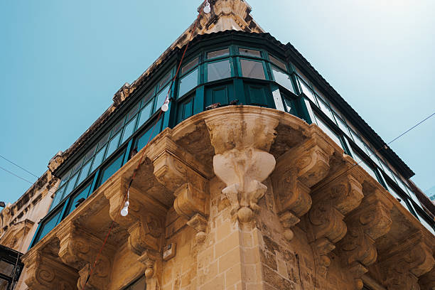 A close-up view of a historic Maltese corner building featuring ornate stone columns, carved capitals, and green-framed windows topped by a bright blue sky and decorative string lights. Malta, Valleta