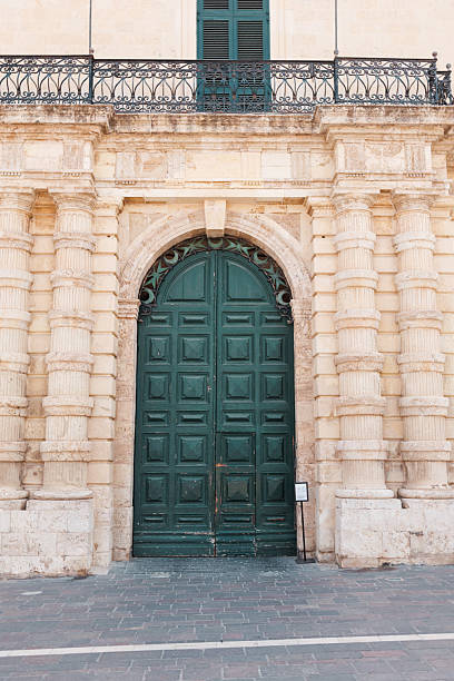 Editorial shot of a large green arched door set in a limestone Maltese façade. Stone columns and an ornate balcony railing convey historic architecture, texture, and Mediterranean charm. Malta, Valleta