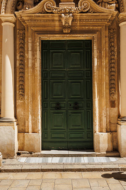 A grand, weathered entrance featuring a dark green paneled door set within an elaborately carved stone arch in Malta. Warm sunlight highlights textured columns and ornate details, conveying historic charm and cultural heritage.