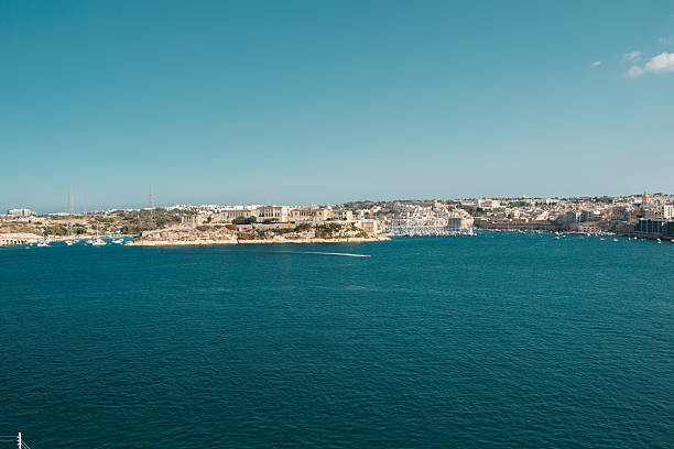 A wide coastal view of a Maltese harbor town showing clear blue sea, historic limestone buildings, and a calm waterfront under a bright sky. Ideal for travel, tourism, and Mediterranean lifestyle themes.