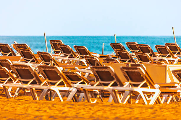 Rows of sunlit beach loungers sit empty on golden sand, framed by blue sea under a cloudless sky. Relaxing scene evokes summer vacation, travel, and leisure at a beautiful seaside destination