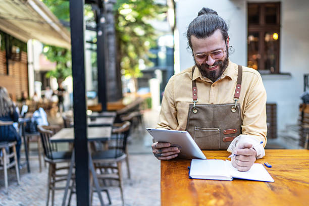 istock Smiling man wearing apron checking tablet and writing in cafe 2237740465