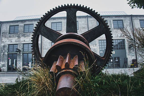 Large rusty industrial gear displayed outside historic factory building — symbol of engineering, machinery, and industrial heritage.