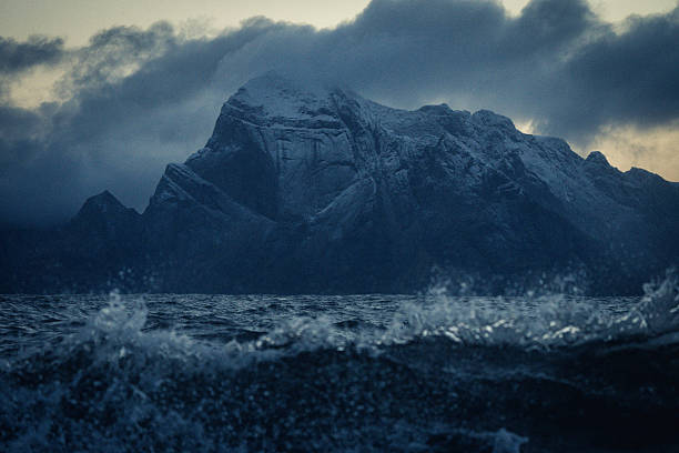 Dramatic view of dark waves crashing beneath snow-covered mountains at twilight, capturing the wild power of Arctic nature.