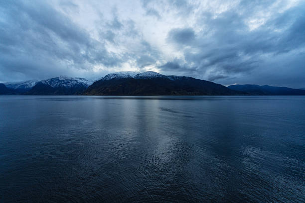 Serene view of a Norwegian fjord surrounded by autumn foliage and distant snow-covered mountains under a moody evening sky.