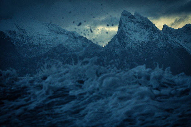 Dramatic view of dark waves crashing beneath snow-covered mountains at twilight, capturing the wild power of Arctic nature.