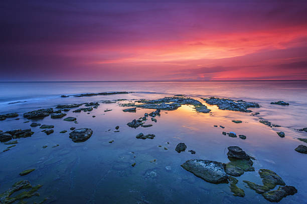 Scattered dark rocks emerge from the glassy Mediterranean waters like natural sculptures, their forms perfectly reflected in the mirror-smooth surface beneath a spectacular twilight sky that transitions from deep purple to brilliant pink and orange.