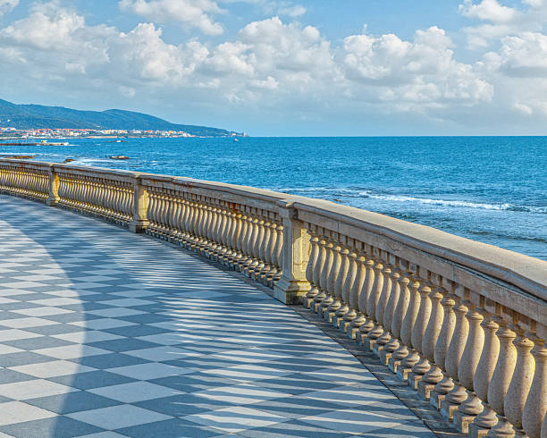 The magnificent Terrazza Mascagni extends gracefully along the Livorno coastline, its ornate neoclassical balustrade with decorative balusters creating elegant shadows across the distinctive black and white checkerboard marble flooring while the brilliant blue Mediterranean Sea stretches toward distant mountains.