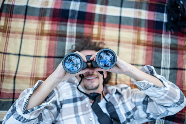 A man relaxes on a plaid blanket outdoors, peering through binoculars. Reflections of trees and sky appear in the lenses, conveying curiosity, exploration, and connection with nature.