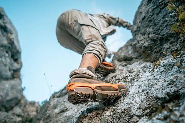 A climber ascends a rocky cliff, wearing rugged hiking boots with crampons for traction. The clear blue sky and natural landscape convey adventure, focus, and outdoor exploration.