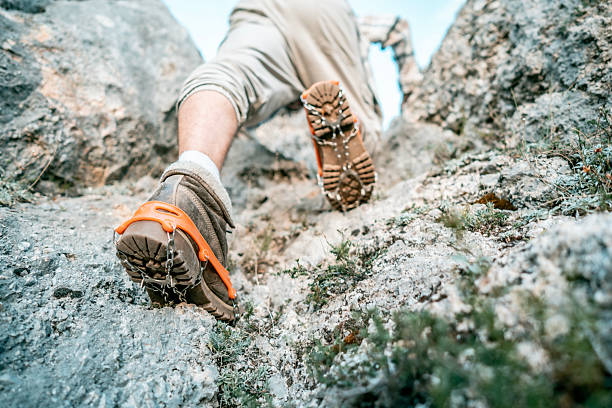 Close-up shot of a hiker's booted feet gripping a rocky trail with rugged terrain and metal traction devices. Emphasizes outdoors, adventure, mountaineering, and determination.