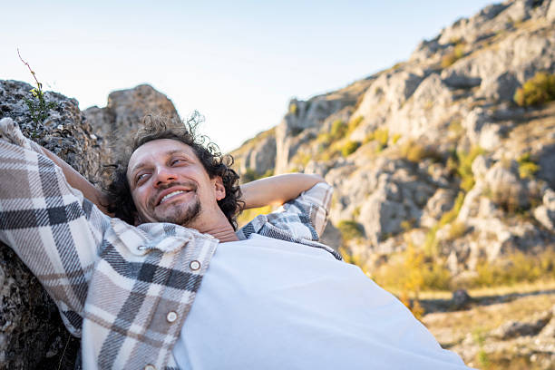 A man in a plaid shirt rests on a rocky surface in a sunlit mountain landscape, smiling as he relaxes. The scene conveys leisure, freedom, and outdoor adventure.