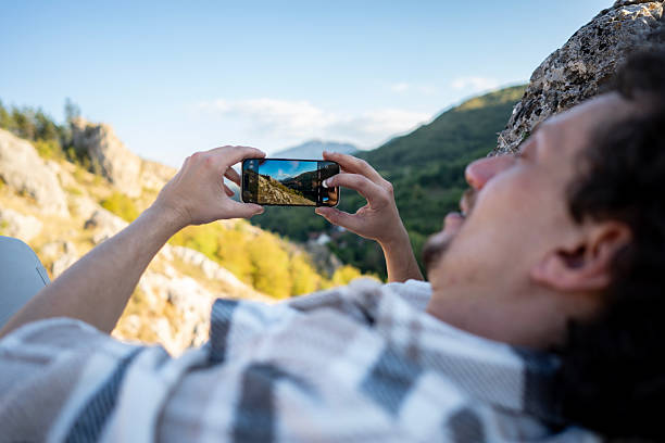 A man relaxes on sunlit rocks while filming a rugged mountain landscape with his smartphone, conveying outdoor leisure, travel vibes, and the joy of capturing nature.