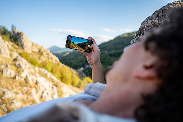 A traveler lies on a rocky ledge, holding a smartphone to photograph a dramatic mountain landscape. Clear blue sky, rugged slopes, and green valleys create a vivid outdoor memory.