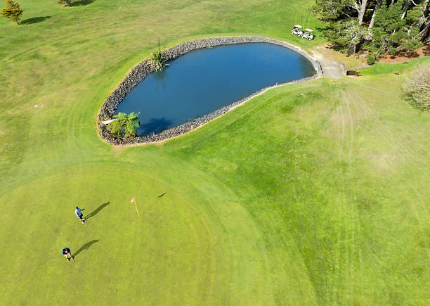 Male golfer swings golf club by a water hazard, with team watching, highlighting the challenge and teamwork on a lush golf course, surrounded by trees.