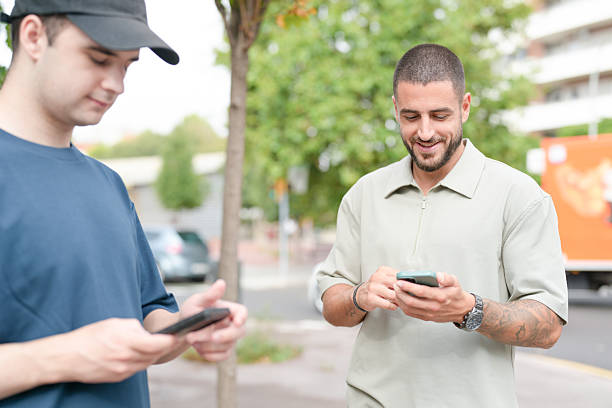 Two men are standing on a sidewalk, both looking at their cell phones. One man is wearing a blue shirt and a hat