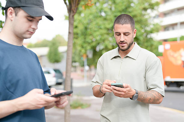 Two men are looking at their cell phones on a sidewalk. One man is wearing a hat. The other man is wearing a blue shirt