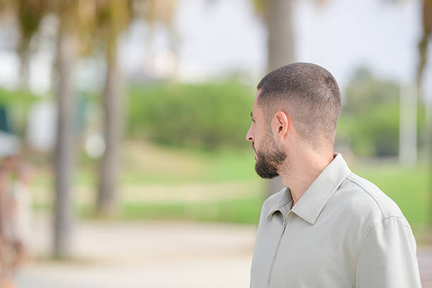 A man with a shaved head and beard is standing in a park. He is wearing a white shirt and looking off into the distance