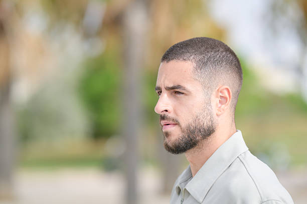 A man with a shaved head and a beard is standing in a park. He is wearing a white shirt and looking at the camera