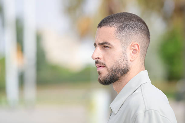 A man with a shaved head and a beard is standing in front of a building. He looks serious and focused