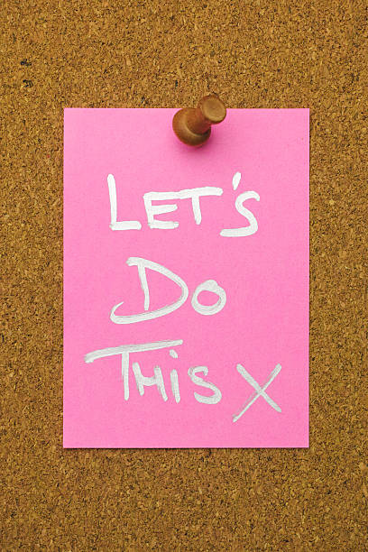 The words 'LET'S DO THIS X' written in a silver marker pen on pink note paper, pinned by a wooden pin to a cork message board. Conceptual motivational and inspirational quote message relating to conquering adversity, together, teamwork, finish, completion, etc.