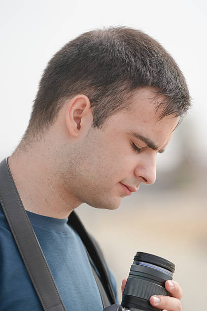 A man with a beard and short hair is holding a camera and a cup. He looks tired and is looking at the camera
