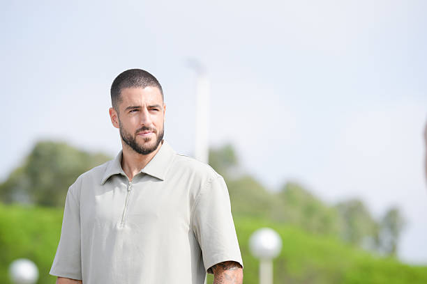A man with a shaved head and a beard is standing in a park. He is wearing a grey shirt and a grey hat