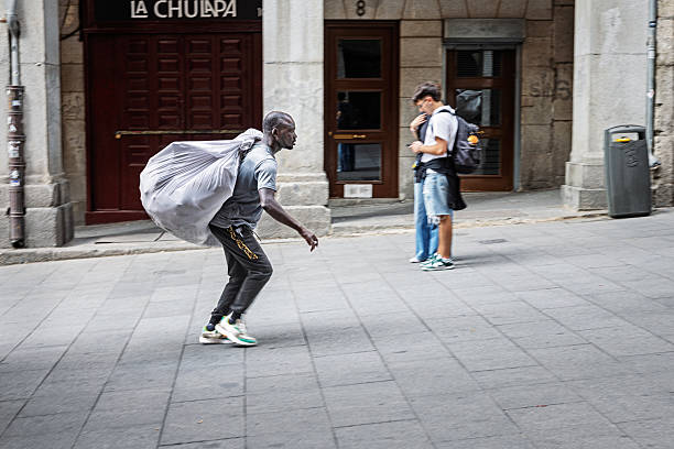 Madrid, Spain, Europe, September 8, 2025:  A street vendor walks briskly through downtown Madrid, Spain, carrying a large sack of merchandise over their shoulder. Dressed in athletic wear, the individual navigates the urban landscape with purpose, reflecting the informal economy and entrepreneurial spirit found in many European cities. In the background, pedestrians linger near a building entrance marked La Chulapa, while graffiti, signage, and a trash bin add texture to the street scene. This editorial image captures the dynamic rhythm of city life, highlighting mobility, labor, and the everyday hustle in the Spanish capital.