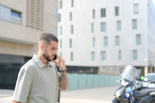 A man is talking on his cell phone while walking down the street. He is wearing a grey shirt and has tattoos on his arm. The scene is set in a city with a motorcycle parked on the side of the road