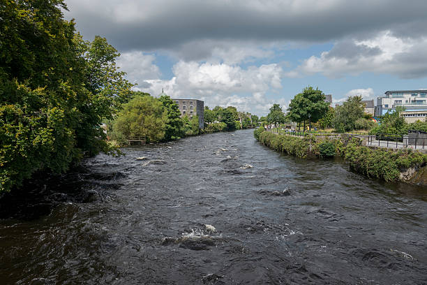 The river Corrib flowing through Galway, Ireland, with modern buildings on one bank and a pathway with lush greenery on the other, under a partly cloudy sky