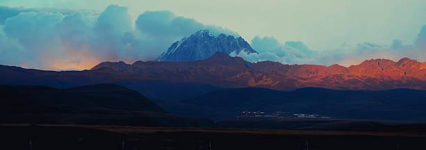 This breathtaking panoramic landscape showcases a towering snow-capped peak piercing through swirling blue clouds, flanked by layered crimson and orange ridges bathed in the warm glow of sunset, descending into a shadowed valley dotted with traditional white-roofed buildings and golden grasslands. The ethereal play of light and mist evokes awe and serenity, ideal for adventure travel, nature inspiration, Himalayan exploration, or motivational visuals in editorial, advertising, or wellness campaigns.