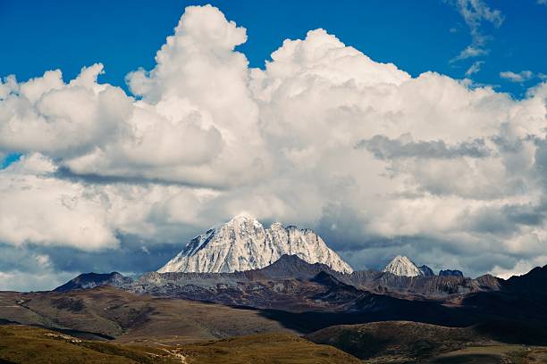 This stunning panoramic landscape features a towering, jagged snow-capped peak dramatically emerging from swirling white clouds under a vast blue sky, with cascading layers of earthy brown ridges and valleys below evoking epic scale and natural wonder. The high-contrast composition and ethereal light capture the raw beauty of alpine wilderness, ideal for adventure travel, nature inspiration, outdoor exploration, or motivational visuals in editorial, advertising, or tourism campaigns.