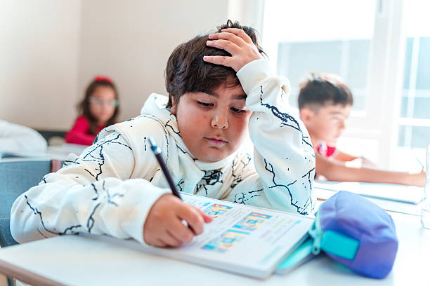 Cheerful chubby-cheeked, dimpled, dark-skinned child  young boy sitting at his desk in a classroom, holding a black pen and writing in a book