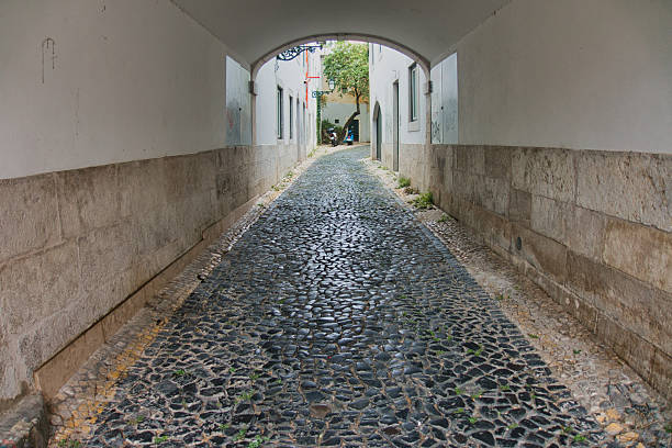 Cobbled Street View through Tunnel Arch - Perspective view down a narrow, empty pedestrian street paved with traditional Portuguese cobblestones, framed by a stone archway.