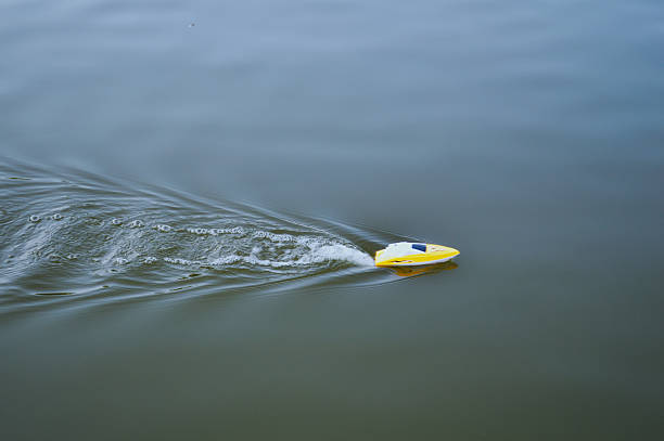 Photo of a small yellow remote-controlled boat creating ripples on a calm water surface while moving. The image captures leisure activity, water recreation, and outdoor fun.