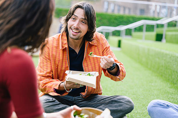 Man in his 20s enjoying picnic with friend outdoors, smiling and listening
