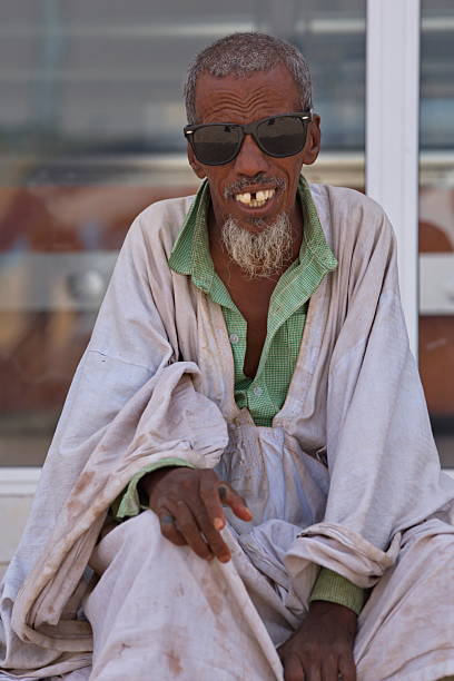 A mid-aged Arabic man wearing a traditional red and white thawb smiles confidently in an outdoor setting. The sunlight adds a warm, welcoming atmosphere to the scene.